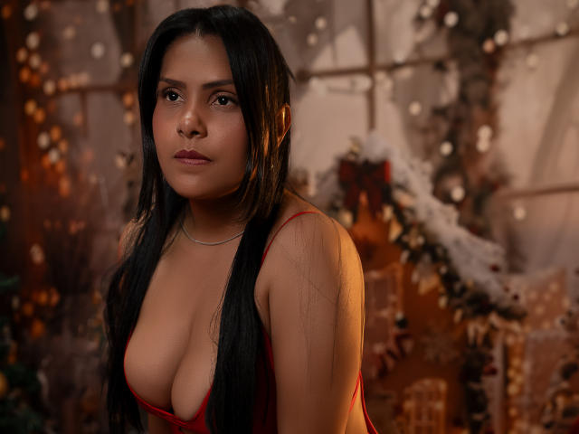 BarbaraSweetty with long black hair, brown eyes, and a red dress showing her big boobs, standing in a warm, golden-lit setting, looking at the camera with a serious expression.