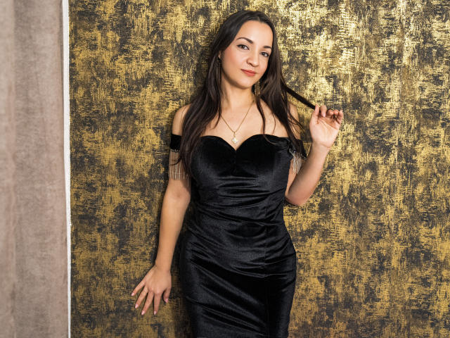 AnyaHappiness stands against a textured gold and black wall. She has long brown hair, slightly wavy, and wears a tight, off-shoulder black velvet dress that highlights her slim figure. Her right hand touches her hair, while her left hand rests on the wall. She wears a simple necklace with a small pendant. The gold texture in the background contrasts with the dark dress, emphasizing her presence. The lighting is soft, highlighting her subtle smile.
