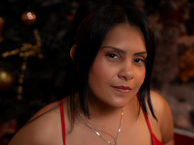 BarbaraSweetty with dark hair, red top, subtle makeup, and a gentle smile, wearing a silver necklace, in a warm-lit, cozy setting. Boobs visible, medium-sized, cute.