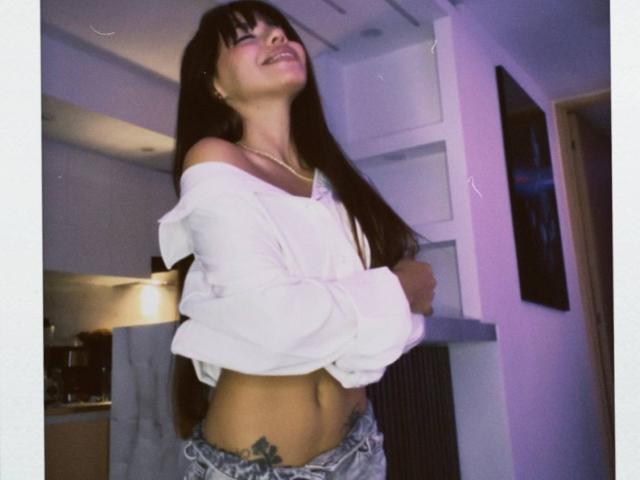 PrincessMiley with long dark hair, smiling, eyes closed, white off-shoulder top, exposing midriff, tattoo on left side, grey pants, standing in modern room, slightly blurred.