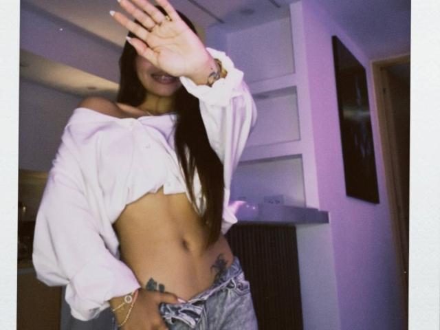 PrincessMiley with long brown hair, white off-shoulder top, and blue jeans, covering her face, showing her toned abs and tattoos on her right hip and lower belly.