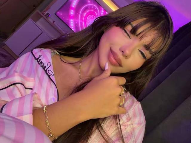 PrincessMiley is in a close-up selfie. She has long brown hair with bangs, a nose ring, and light pink lipstick. She's wearing a pink and white striped pajama top with "tommy" embroidered on it. She has a gold bracelet on her left wrist and a gold ring on her right hand. Her eyes are closed and she's touching her chin with her right hand. The background has a purple neon light and a TV screen with a pink spiral pattern. The image has a warm, purple-tinged light.