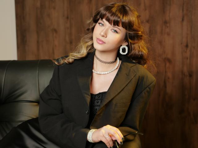 GentleTease is a woman with wavy brown hair and bangs, sitting on a black leather couch. She wears a black blazer over a black, sequined dress, with a diamond choker and matching earrings. Her right hand rests on her lap, showing a ring. Her light skin contrasts with her dark hair. The image has a warm, slightly blurred background. The composition is centered, with GentleTease looking slightly to the right.