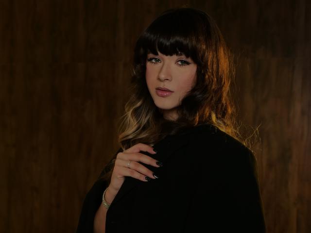 GentleTease is a portrait of a woman with dark brown, wavy hair and blunt bangs. She has light skin, green eyes, and pink lips. She's wearing a black garment and has a silver ring on her left hand, which rests on her chest. The light from the right casts warm shadows on her face and hair. Her nails are painted dark. The texture of the background is wooden. The overall composition is dark and moody.