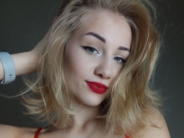 LunaBell has blonde hair, blue eyes, and red lipstick. She's wearing a white wristband, with her hand in her hair, looking at the camera with a slight smile.