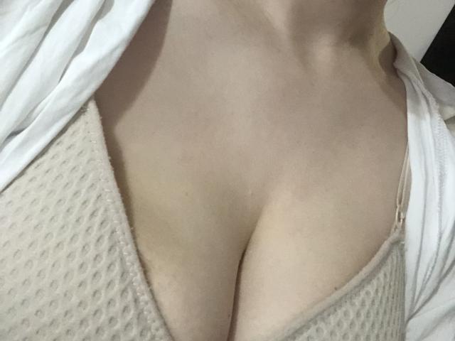 La poitrine pâle de Pelerin, avec son décolleté prononcé, est couverte d'un soutien-gorge beige à pois, et une chemise blanche la recouvre partiellement. Sa peau est lisse et ses seins sont bien visibles..
