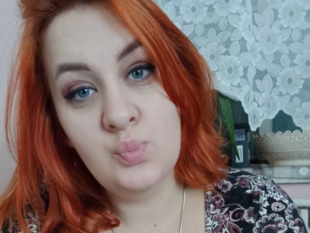 MaryJoysy with orange hair, blue eyes, and pouty lips, wearing a floral top, looking into the camera. Her tits are small, and she's making a silly face.