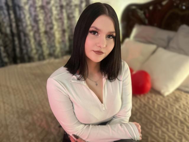 AmeliaGracee with straight black hair, brown eyes, and a nose ring, wearing a white top, smiling softly, her tits slightly visible, in a blurry bedroom.