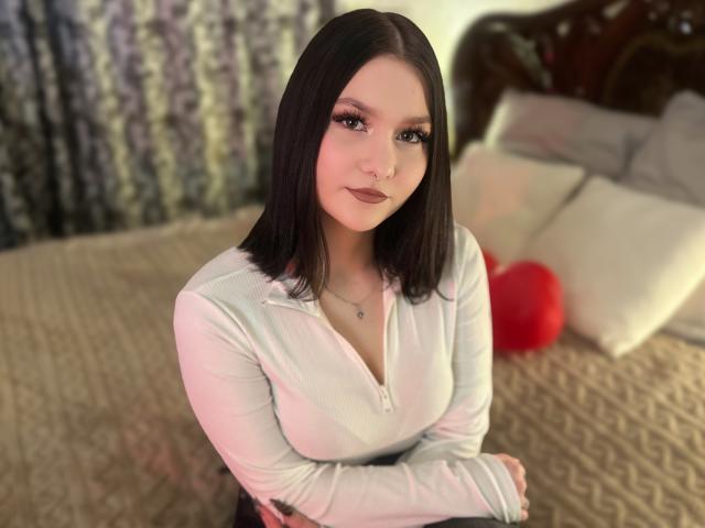 AmeliaGracee with dark straight hair, white v-neck shirt, crossed arms, subtle makeup, nose ring, and small necklace, sitting on a bed with a red heart pillow.