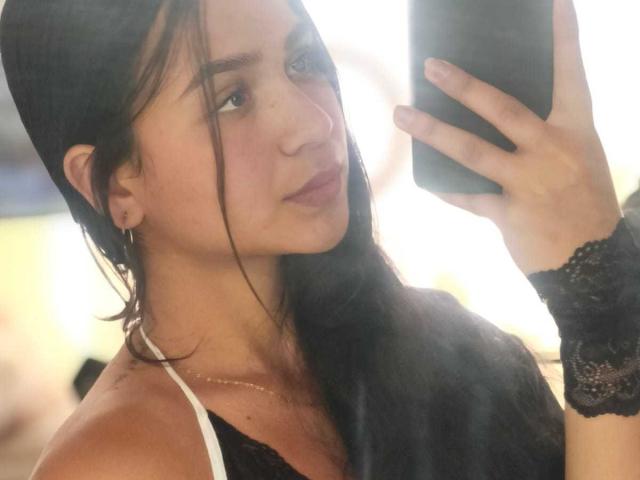 RouxiMori with long black hair, wearing a black lace top and wristband, holding a phone, looking at it, in a mirror selfie, showing her tits and a slight smile.