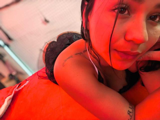 RouxiMori with red lighting, lying on red bed, wearing black lace top, showing tattoo on left shoulder, black hair, looking at camera, hand on cheek.