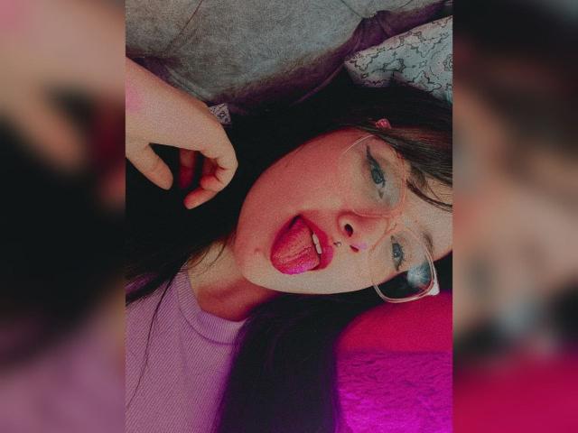 NataliaRivas with dark hair, glasses, and pink lipstick, tongue out, lying down, wearing a purple top, hand near face, blurred faces on sides, colorful, soft lighting.