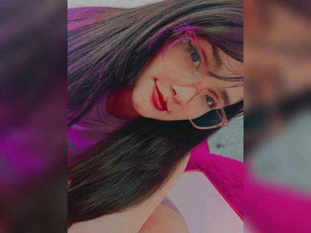 Close-up of NataliaRivas with long, straight, dark brown hair and bangs. She wears pink-tinted glasses and has a small facial piercing below her right nostril. Her light skin contrasts with her dark hair and pink glasses. She has a slight smile with red lipstick. The background includes a pink pillow and a blurred, out-of-focus pink and gray area. The image has a soft, digital filter effect. Her glasses are slightly oversized, adding a playful touch.