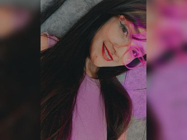 NataliaRivas with long black hair, pink glasses, red lipstick, and pink top, lying down with a cute smile. Her nose is pierced. Her tits are small and she's attractive.