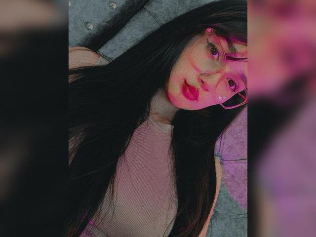 NataliaRivas with long black hair, pink glasses, and pink lipstick, wearing a pink top, lying on a grey tufted surface, with a blurred pink object in the foreground.