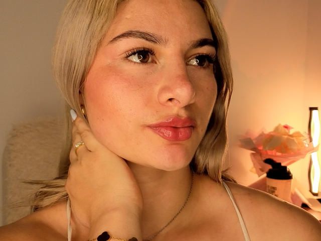 AtheneaRozzi with blonde hair, pink lips, and fair skin, holding her neck, wearing a white top, in a softly lit room with a lamp and pink flowers.