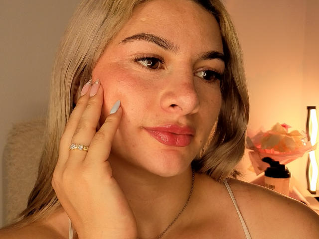 AtheneaRozzi has light brown, shoulder-length hair and a fair complexion. She has brown eyes, slightly blushed cheeks, and full, pink lips. Her right hand, with light blue nail polish and a gold ring, touches her cheek. She wears a thin, beige spaghetti strap. The soft, warm lighting highlights her facial features and creates gentle shadows. A pink floral arrangement and a lit lamp are in the blurred background. The overall composition is centered and focuses on her face. The texture of her skin is smooth. The image has a casual, intimate feel.