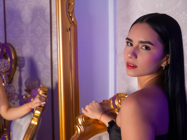 EmmaLeone with long black hair, red lipstick, and gold earrings, looking over her shoulder at a mirror, showing her bare shoulders and a small black top.