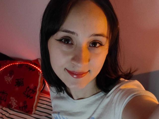 Close-up photo of AnnieLamberth with medium-length black hair, fair skin, and dark, slightly arched eyebrows. She has thick, black eyeliner with a slight cat-eye effect and glossy pink lips. She's wearing a white t-shirt. The background includes a red pillow with black floral patterns and a striped pillow. Warm, reddish lighting casts a subtle glow on her face. Her head is slightly tilted, and she's looking directly at the camera with a slight, confident smile. The image has a casual, intimate feel.