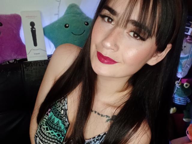 EllieBonneth has dark straight hair, brown eyes, and red lipstick. She's wearing a patterned top, showing a tattoo below her tits. She has a nose ring and eyeliner.