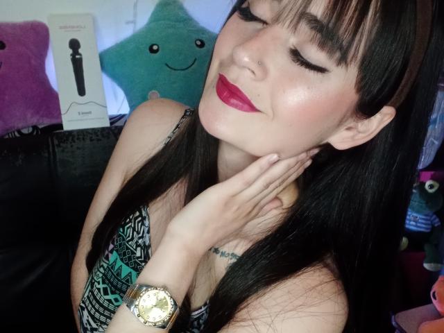 EllieBonneth with closed eyes, dark lipstick, and long black hair, smiling, holding her chin, wearing a watch, and a patterned top, showing a small tattoo on her chest.