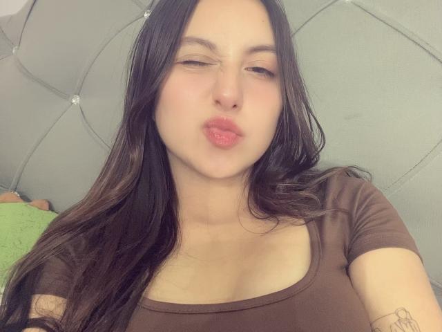 RosalieDupont with long dark hair, pouty pink lips, brown top, tattoo on arm, slightly squinting eyes, light skin, and soft facial expression. Boobs visible under the top.