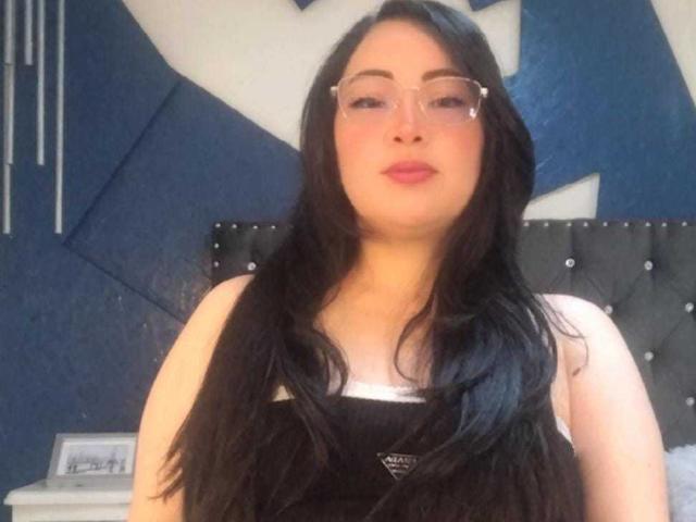 IvyTowen with long black hair, wearing glasses, and a black tank top, has pink lipstick and a neutral expression, standing in front of a blue and black wall.