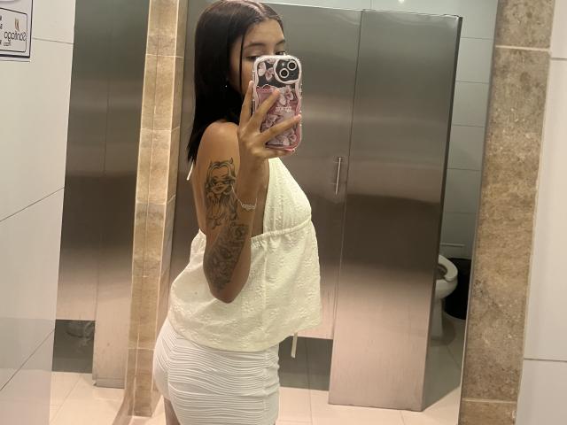 CaritoHam with long dark hair, wearing a white lace top and tight white skirt, showing her tattoos, holding a phone with a pink case, reflecting in a mirror.