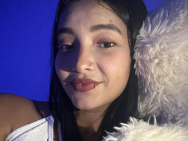 CaritoHam's face with dark hair, brown eyes, and nose ring, wearing a white top, pouting with pink lips, next to a fluffy white pillow, under blue light.