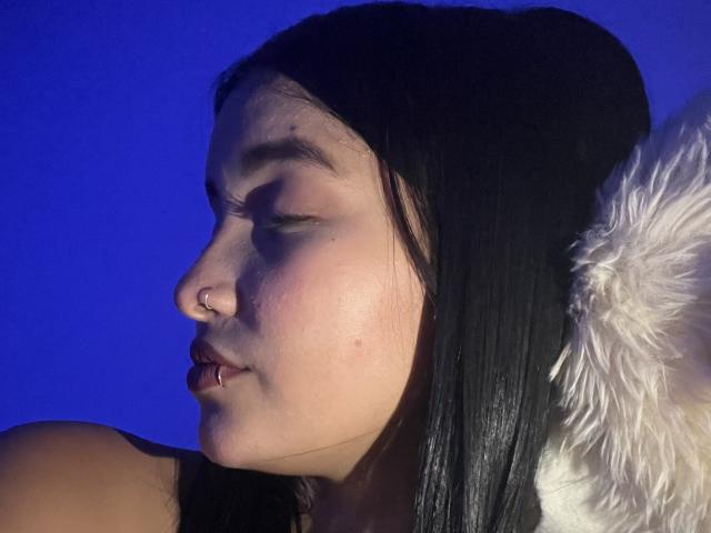 CaritoHam with black hair, nose ring, and pierced lip, eyes closed, blue background, wearing black beanie, white furry shoulder, soft lighting, slight shadow.
