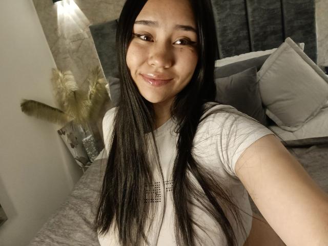 Karollteyes with long black hair, small tits, and a smile in a white tee, taking a selfie in a cozy bedroom. Her cute face and shiny lips are prominent.