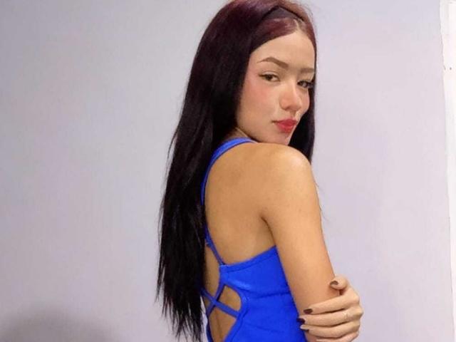 IvorySelene has long, straight, dark brown hair, looking over her shoulder with a slight smile. She wears a bright blue, strappy dress that reveals her back. Her right arm is crossed over her chest, with dark purple nail polish. Her light skin contrasts with the blue dress. The image focuses on her upper body, highlighting her slender build and confident pose. She has subtle makeup, with red lipstick standing out. The overall composition is simple, with IvorySelene centered against a plain background.