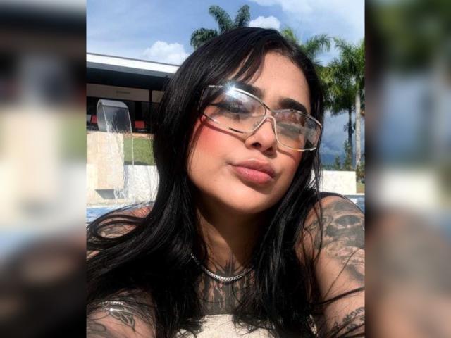 NikkiHill with long black hair, wearing clear glasses, has tattoos on her arms and chest, and is pouting her pinkish lips. She's outside in bright sunlight.