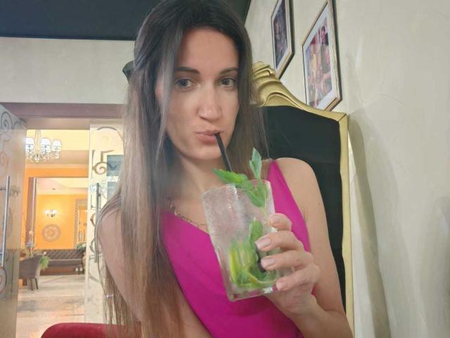 BanyReid with long brown hair, pink dress, sucking on a drink with a straw, holding a glass with green leaves, pouty face, and a small nose ring.