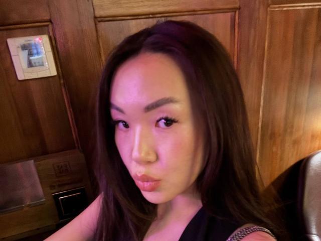 QueenxBae has long brown hair, full lips, and a cute face with makeup, looking at the camera with a slight pout, sitting in a wooden room.