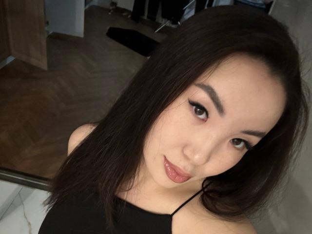 QueenxBae has shiny black hair, dark eyebrows, and glossy pink lips. She's wearing a black top, looking at the camera with a slight smile. Her tits are not visible.