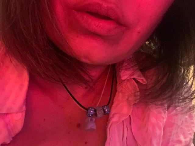 ElizavetaCream's pink-lit face showing parted lips, wearing a white shirt with a necklace featuring two blue beads and a small bell, brown hair partially covering her mouth.