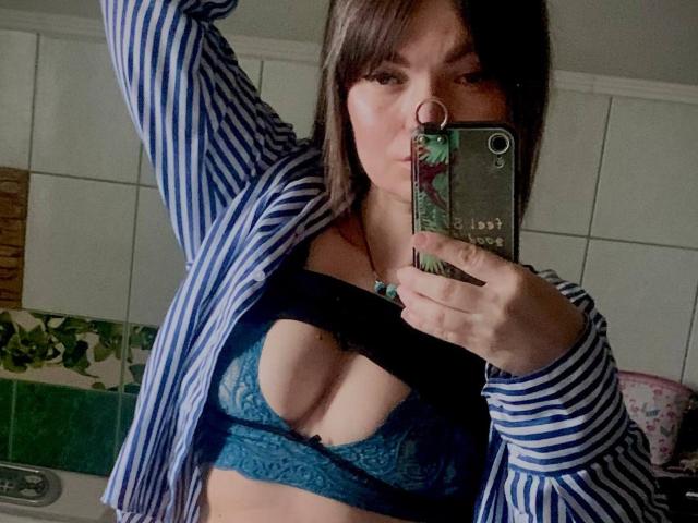 ElizavetaCream with brown hair, striped shirt open, blue lace bra showing her tits, arm raised, taking a mirror selfie with a green phone.