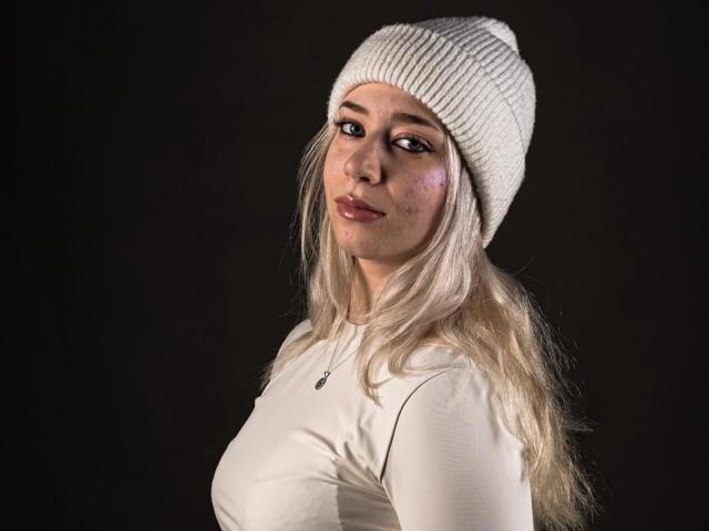BeckyThaissa with long blonde hair, white knit beanie, and white long-sleeve shirt, showing her boobs slightly, looking to the side with a subtle smile, against a dark background.