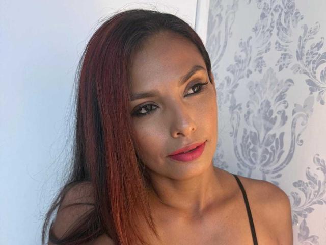 RoxaneMoore with red and black hair, brown eyes, and pink lipstick, wearing a black strap, looking at the camera with a slight smile. Her tits are not visible.
