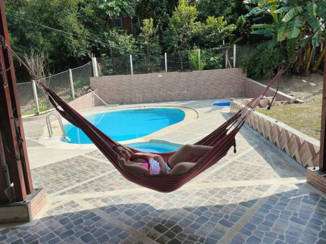 A hammock with dark red stripes hangs in a sunny backyard, holding ElyinRojas who has a pink bikini top and brown skin. ElyinRojas' legs are bent at the knees, feet up. A small, blue-tiled swimming pool is in the background, surrounded by a stone-tiled patio and a low brick wall. Green trees and a tall banana plant are behind the wall. The hammock is suspended from a wooden frame on the left. The image uses bright natural light.