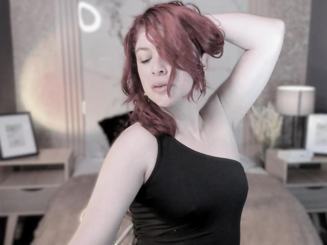 MoonXVelvet with pink hair, black one-shoulder top, arm raised, eyes closed, slight pout, soft light, subtle makeup, bedroom setting, medium shot, relaxed, sexy, confident.