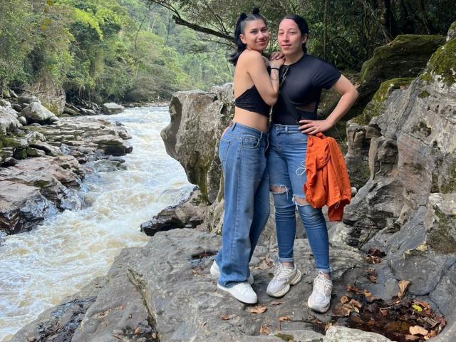 Two HotHarmonyX standing on rocks by a flowing river. Both have black tops, blue ripped jeans, and white sneakers. One holds an orange shirt. Both have small tits.