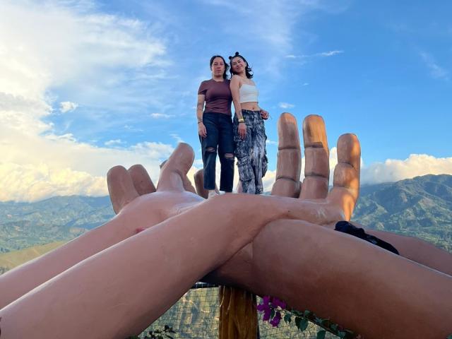 Two HotHarmonyX standing on a pair of outstretched hands, blue sky above, mountains in background. One wears a maroon top, other in a white top, with patterned pants.