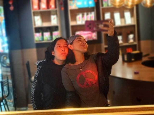 HotHarmonyX with black hair, black hoodie, and HotHarmonyX with dark hair, bandana, gray shirt, taking selfie in cafe, smiling, looking up, mirror reflection, pink phone.