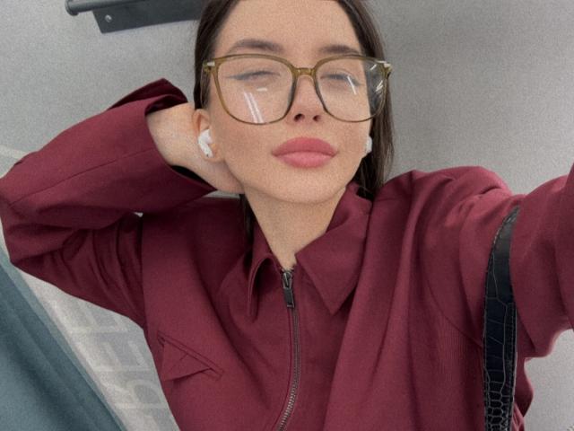 JeanieCat with brown glasses, pink lipstick, and white earbuds, wearing a maroon zip-up jacket, hand in hair, slightly smug expression, medium tits, straight brown hair, casual selfie.