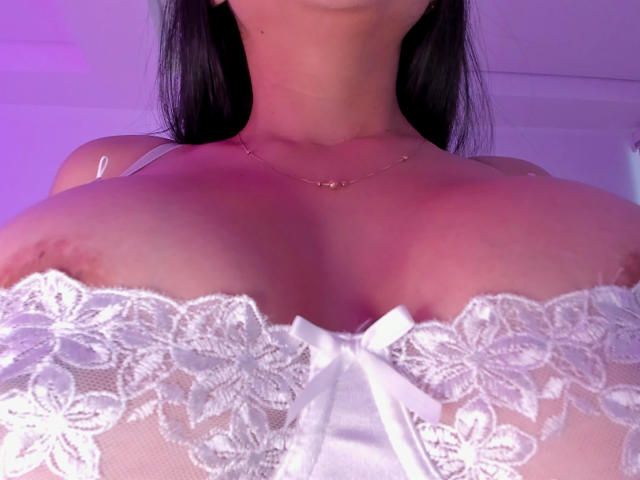 VictoriaGiulias avec de grandes seins, portant un soutien-gorge en dentelle blanche avec des motifs floraux et un petit nœud, sa poitrine et son cou visibles, cheveux foncés, un collier en or discret, éclairage violet..