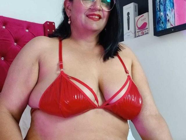 RubyRosh, plus-sized woman with red, shiny vinyl bra, showing large, shiny tits, smiling, wearing glasses, black hair, pink headboard, white wall, colorful artwork, and sex toy box.