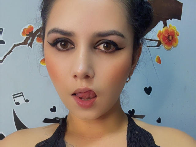 Kkatalina has dark eyes, thick black eyeliner, and is sticking out her pink tongue. She's wearing a black halter top and has her hair in a bun.