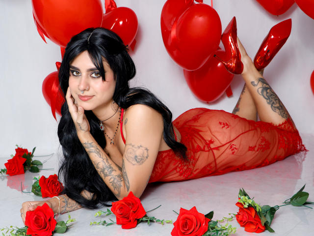SamanthaGio with black hair, red lace dress, tattoos, red high heels, lying on red roses, red balloons, and red roses, looking at camera, smiling, with hand on cheek.